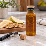 Open Honey Feast Wildflower Honey 8oz Muth glass jar with its removed tapered cork on a wine corkscrew, beside cheese, crackers and pear.