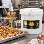 Honey Feast Allergy Blend Honey 2-gallon bucket beside a stainless steel sheet pan of granola with honey being poured from a ladle.