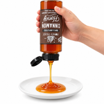 Hand squeezing an inverted Honey Feast Cinnamon Honey 12oz plastic bottle onto a white plate with the flip-top lid open and label correctly upside down