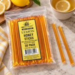 Honey Feast Lemon Honey Sticks 50-pack lying flat beside lemon tea and fresh lemons, with full-length loose honey sticks