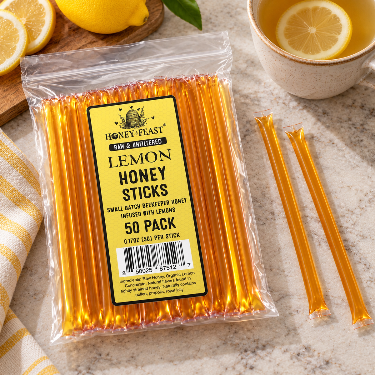 Honey Feast Lemon Honey Sticks 50-pack lying flat beside lemon tea and fresh lemons, with full-length loose honey sticks