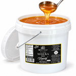 Open Honey Feast Manuka Infused Honey with Propolis 2-gallon bucket, with honey pouring from a stainless steel ladle on a white background.