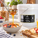 Honey Feast Manuka Infused Honey with Propolis 2-gallon bucket beside a breakfast bowl with fruit and granola, with honey poured from a stainless steel ladle.