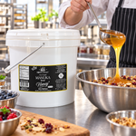 Honey Feast Manuka Infused Honey with Propolis 2-gallon bucket in a commercial kitchen as a cook ladles honey into a stainless steel mixing bowl.