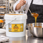 Honey Feast Orange Blossom Honey 2-gallon bucket beside a cook ladling honey into a stainless steel mixing bowl in a commercial kitchen.