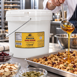 Honey Feast Orange Blossom Honey 2-gallon bucket beside a stainless steel sheet pan of granola with honey being poured from a ladle.