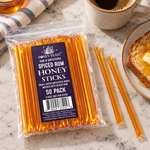 Honey Feast Spiced Rum Honey Sticks 50-pack lying beside coffee and buttered toast, with loose honey sticks
