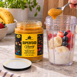 Adding a spoonful of Honey Feast Superfood Honey to a blender cup with fruit beside the 12oz glass jar