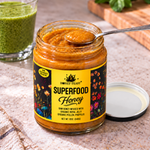 Spoonful of thick Honey Feast Superfood Honey above an open 12oz glass jar beside a green smoothie