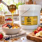 Honey Feast Black Gum Tupelo Honey 2-gallon bucket beside a breakfast bowl with fruit and granola, with honey poured from a stainless steel ladle.
