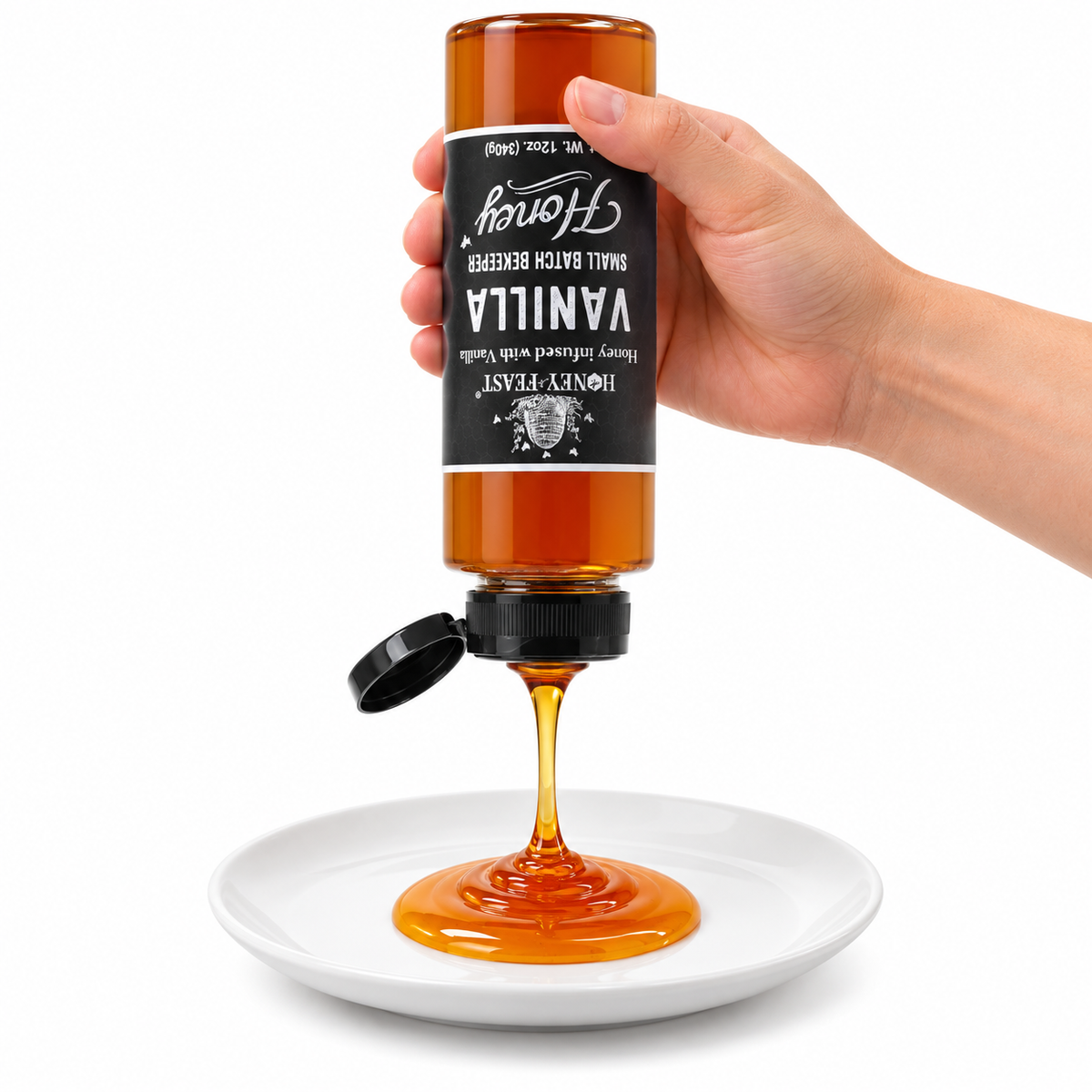 Hand squeezing an inverted Vanilla Honey 12oz plastic bottle onto a white plate with the flip-top lid open and label correctly upside down