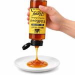 Hand squeezing an inverted Honey Feast Wildflower Honey 12oz plastic bottle onto a small white plate, with the original yellow label correctly upside down